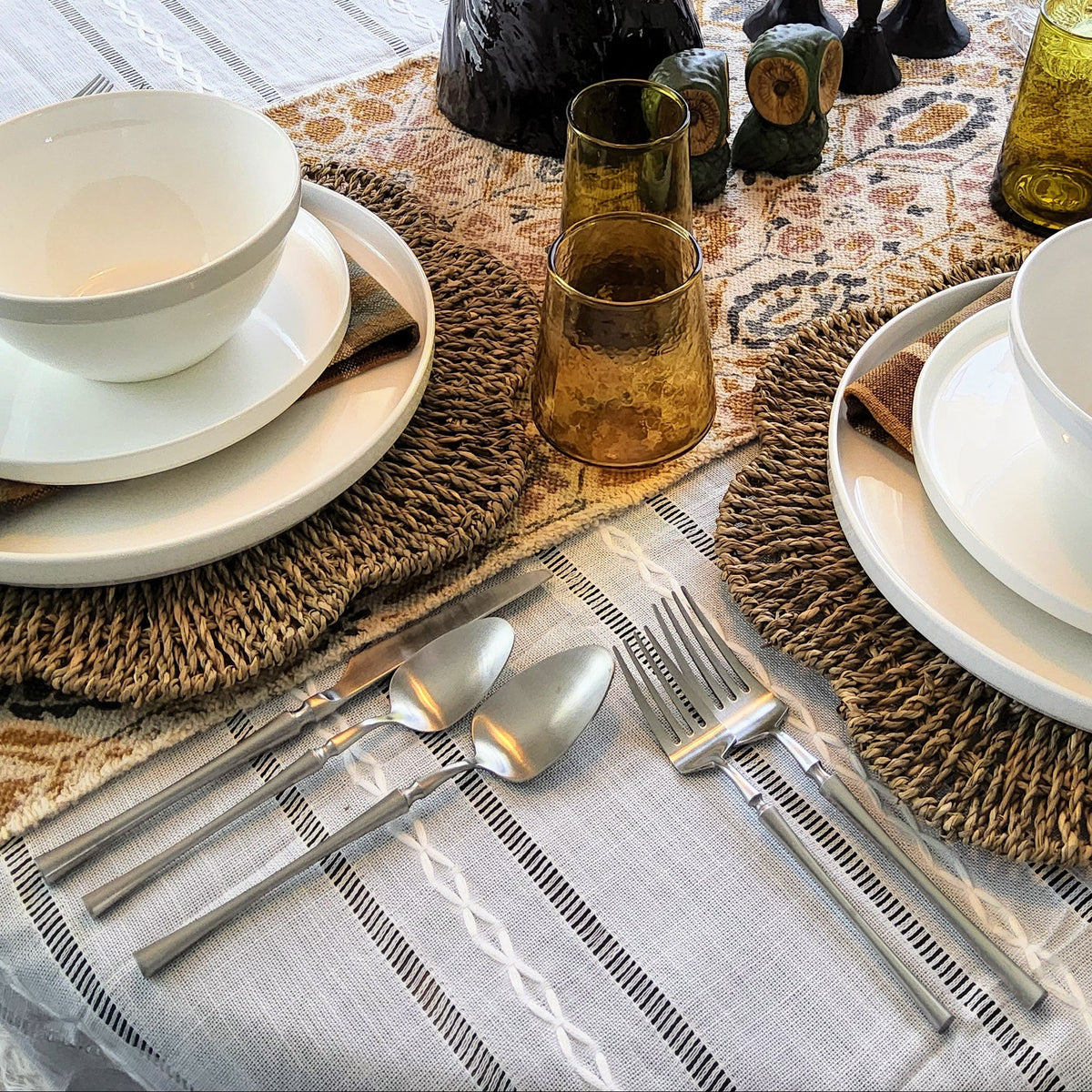 A styled round dining table set for two, featuring white ceramic bowls and plates, silver cutlery, woven placemats, plaid napkins, amber glass tumblers, and a central floral centerpiece on a patterned table runner.