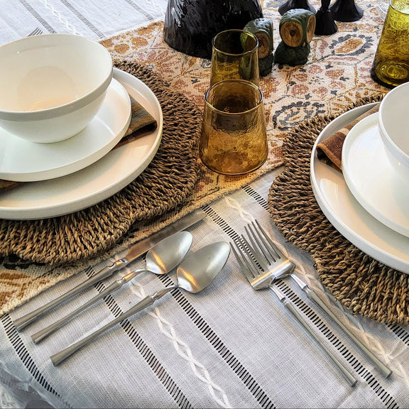 A styled round dining table set for two, featuring white ceramic bowls and plates, silver cutlery, woven placemats, plaid napkins, amber glass tumblers, and a central floral centerpiece on a patterned table runner.
