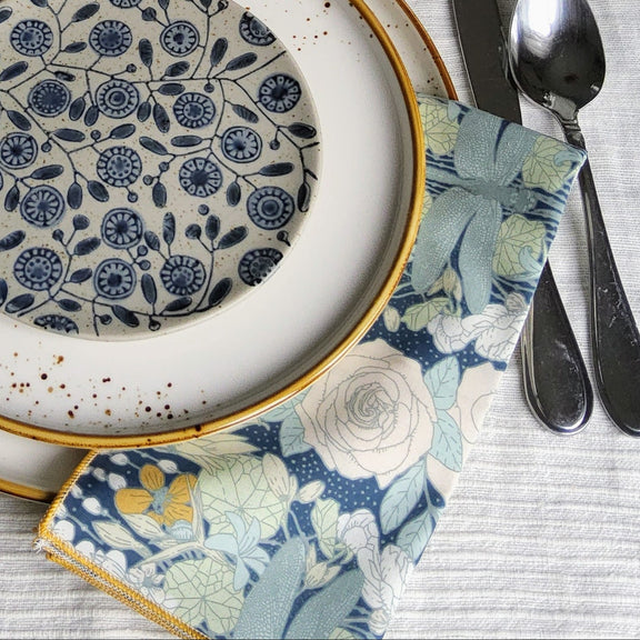 A styled table setting featuring a place setting with a white ceramic charger plate, a smaller white plate with gold rim, a blue and white floral patterned plate, a matching small bowl, a floral napkin, and silver cutlery on a textured white tablecloth.