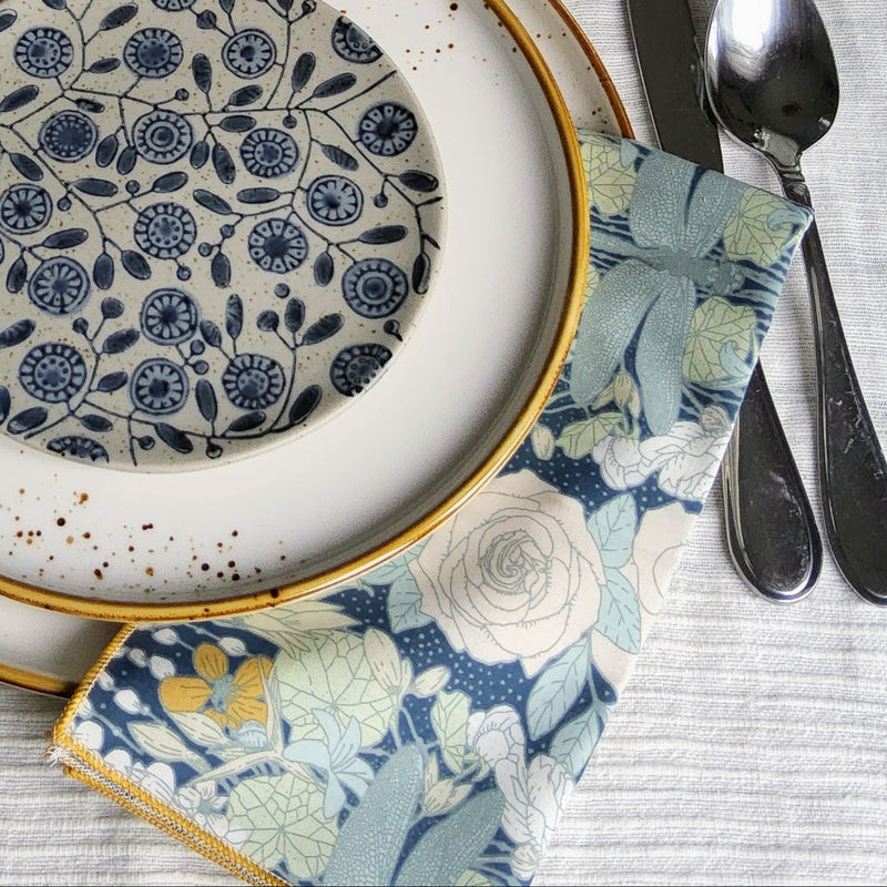 A styled table setting featuring a place setting with a white ceramic charger plate, a smaller white plate with gold rim, a blue and white floral patterned plate, a matching small bowl, a floral napkin, and silver cutlery on a textured white tablecloth.