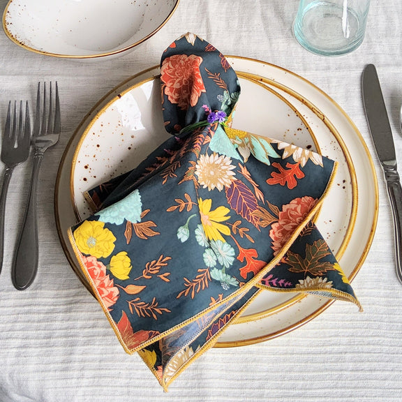 A folded floral napkin with a dark navy background and multicolor botanical print, styled on a gold-rimmed plate in a tabletop setting with cutlery and a glass.